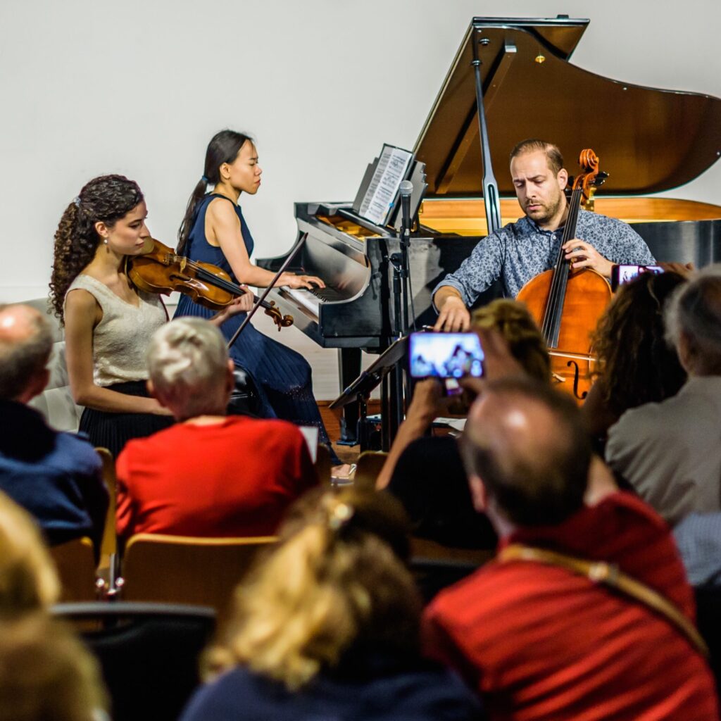 A violinist, pianist, and cellist perform for an audience with mixed ages, including older adults