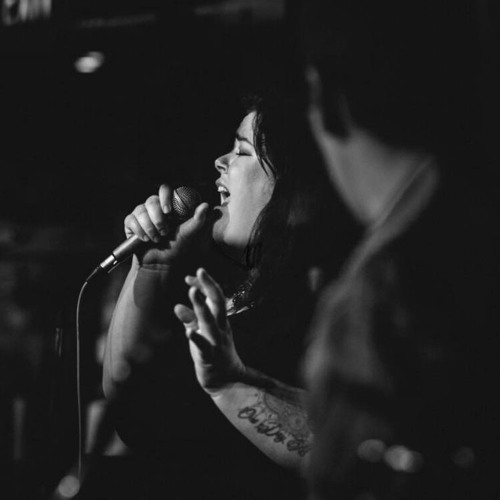 A black and white photo of Jenna Marie Pinard singing onstage and holding a microphone. There is a guitarist to the front right of her in the frame.