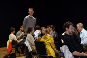 Groups of people sit in chairs in circles while a man stands and speaks
