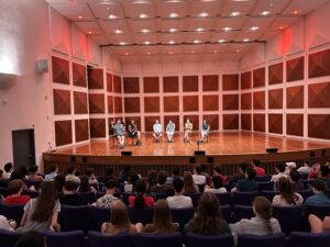 A group of six people presents to a large audience in an auditorium