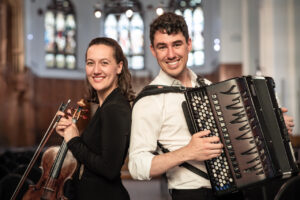 Violinist Yolanda Bruno, and accordionist Michael Bridge post back to back. They are both holding their instruments and smiling toward the camera.