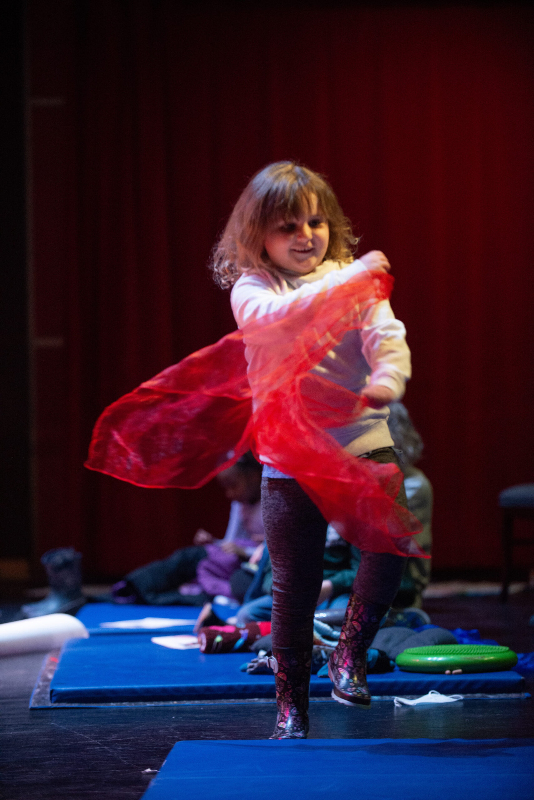 A young girl wearing rubber boots dances with a red scarf in a concert hall