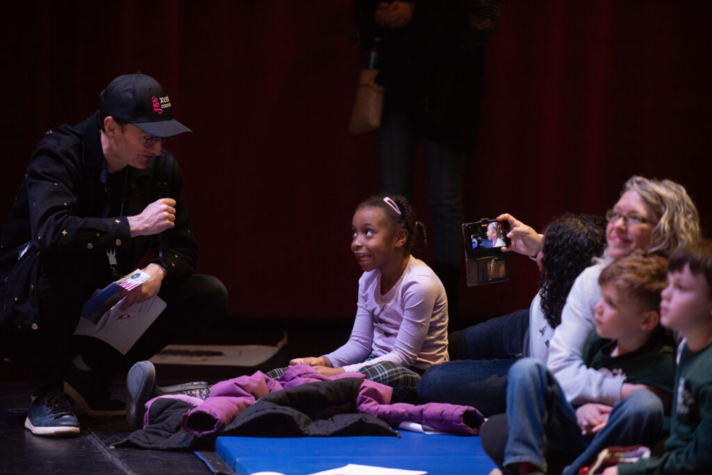 A girl sits on the floor looking up at a man who is crouching down with a microphone.