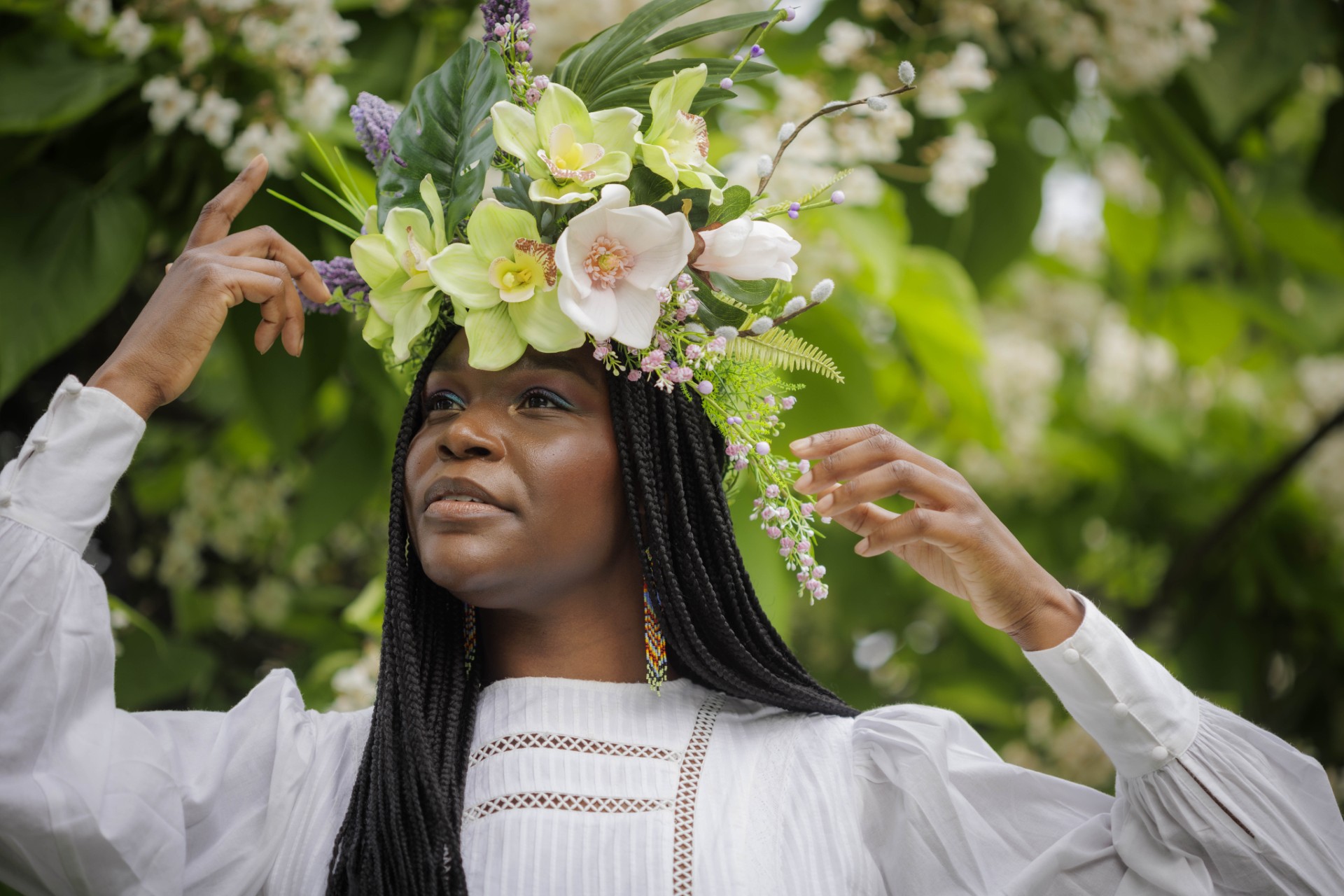 A woman with long braids holds her hands above her shoulders, wearing a white dress and a flower headdress.